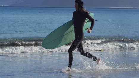 Video-of-caucasian-man-with-dreadlocks-in-wetsuit-carrying-surfboard-running-in-sea-on-sunny-beach