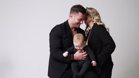 caucasian couple in love wear black jackets and kissing tenderly. beautiful portrait of family of three in studio. white backdrop.