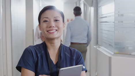 portrait of female nurse wearing scrubs with digital tablet in busy hospital corridor