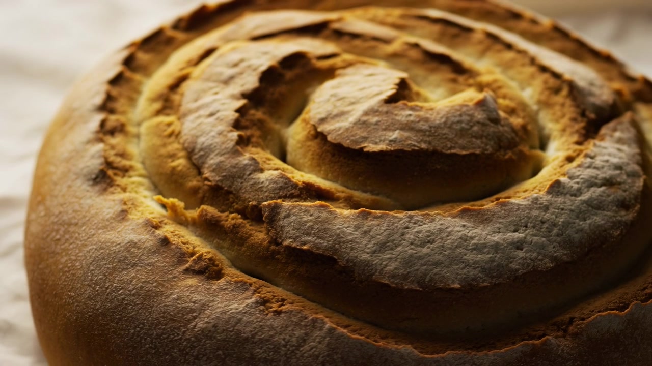 Una pagnotta di pane artigianale rotonda riempie una scena morbida e sfocata, mostrando incisioni a spirale, creste screpolate, crosta dorata, spolverata di farina e consistenza cotta su un tavolo rustico di stoffa, con pane, pagnotta, crosta, spirale.