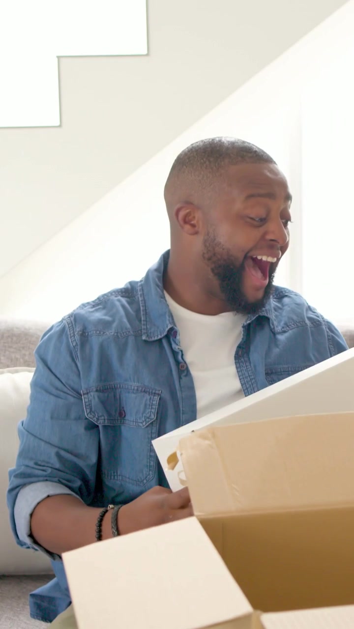 Vertical video: Opening delivery box man in denim lifting white box on sofa removing blue tissue