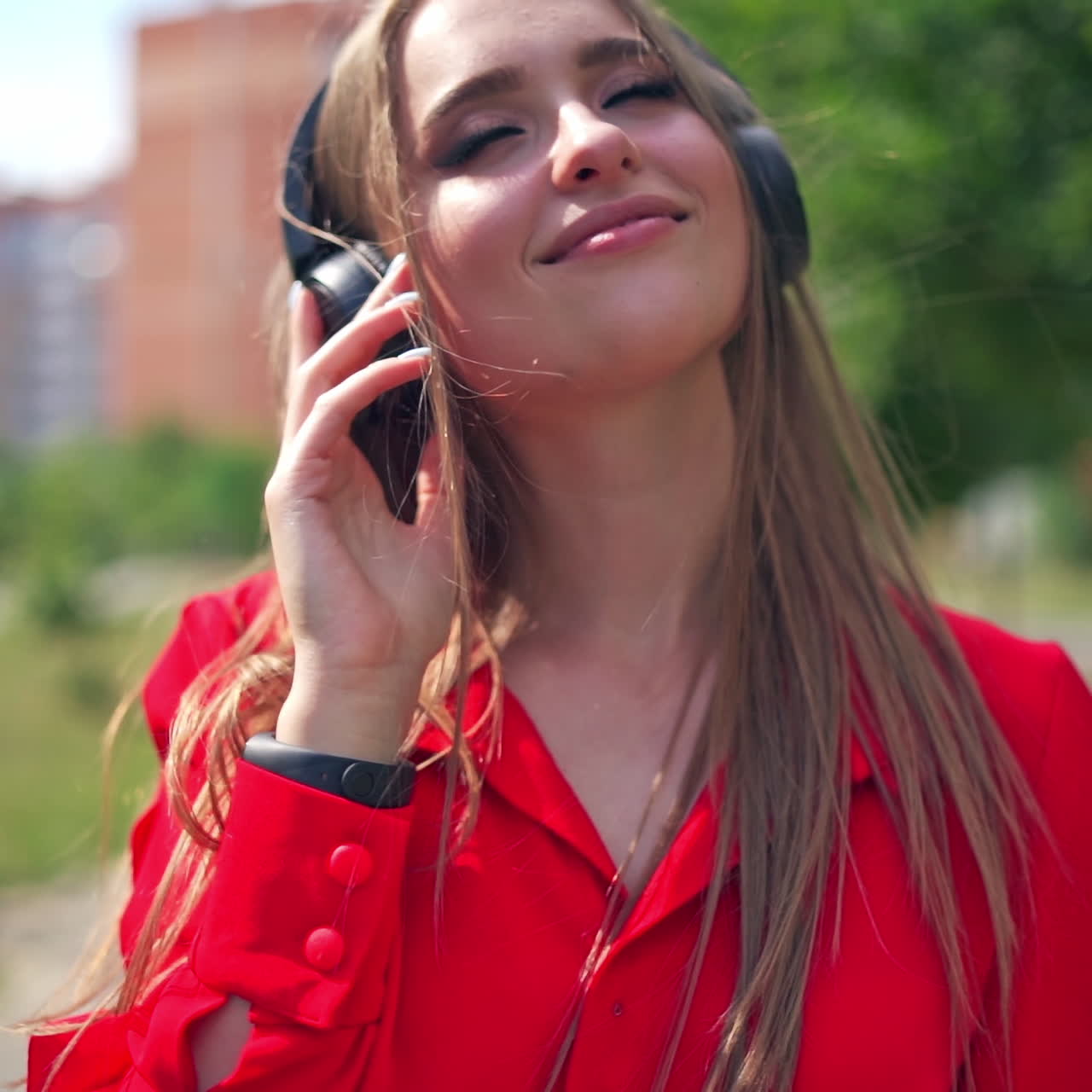 Attractive woman listening to music outdoors. Young female in red dress with headphones enjoys music in the city street in summer.