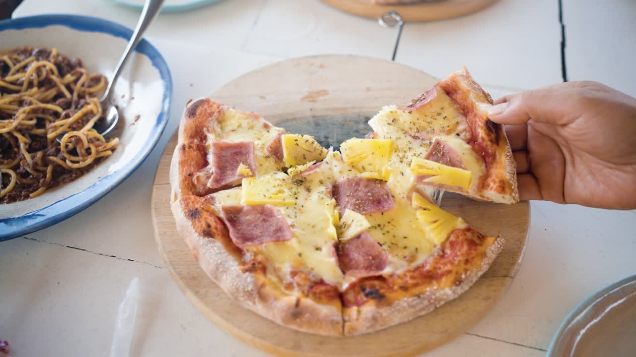 Primo piano al rallentatore di persone che condividono una pizza hawaiana con formaggio fuso filante, prosciutto e ananas, su un tagliere di legno a un tavolo da pranzo informale in vacanza.