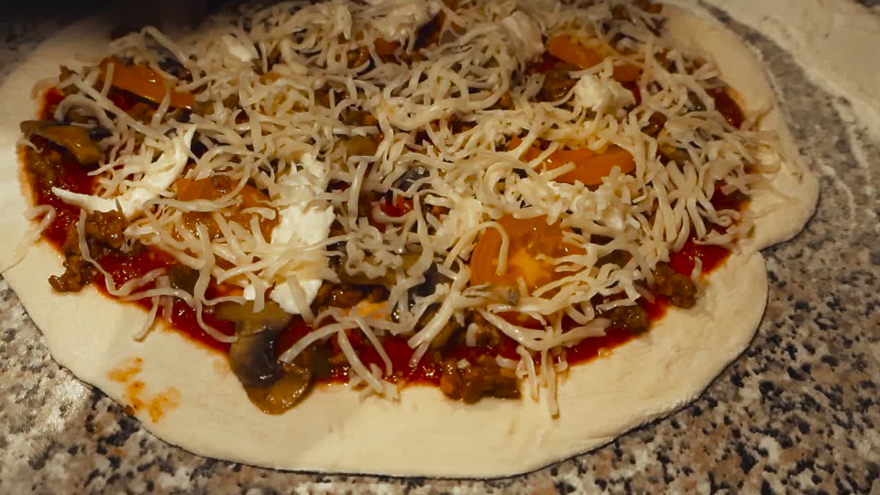 Closeup orbiting of chef hand putting shredded cheese over handmade pizza base covered with vegetables ingredients, meat toppings and tomato sauce in slow motion. Low angle fresh dough on home kitchen