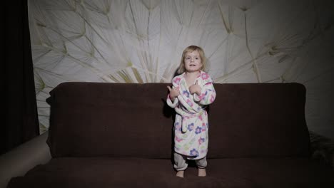 Little-cute-girl-with-blond-hair-jumps-on-sofa.-Bathrobe-clothes.-Happy