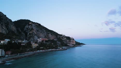 Menton-near-the-italian-border-during-twilight,-showcasing-the-serene-mediterranean-coast-and-the-town-nestled-against-rugged-hills,-aerial-view