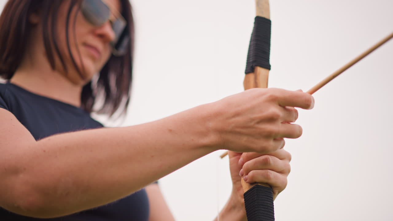 white woman archer preparing traditional bow in open field, close detail on hands drawing string and seating arrow, leather grip and rugged posture suggesting lone warrior training, focused gaze