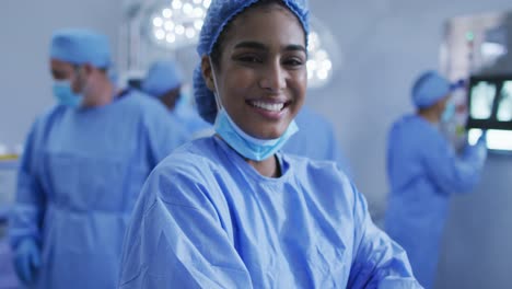 Portrait-of-mixed-race-female-doctor-standing-in-operating-theatre-smiling-to-camera