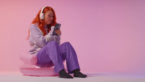 studio shot of young gen z woman wearing headphones streaming music to mobile phone 4