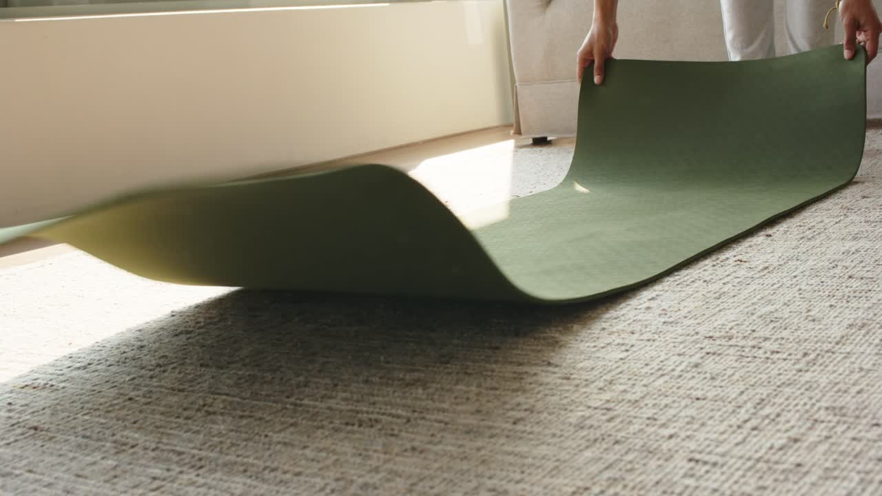 Person i treningsklær strekker seg og griper en rullet grønn matte som rulles ut på et beige teppe for trening