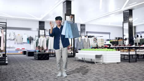 옷 가게 에 서 있는 동안 쇼핑 가방 을 들고 손 을 흔들고 카메라 에 미소 짓고 있는 아시아 의 쇼핑 남자 의 전체 모습