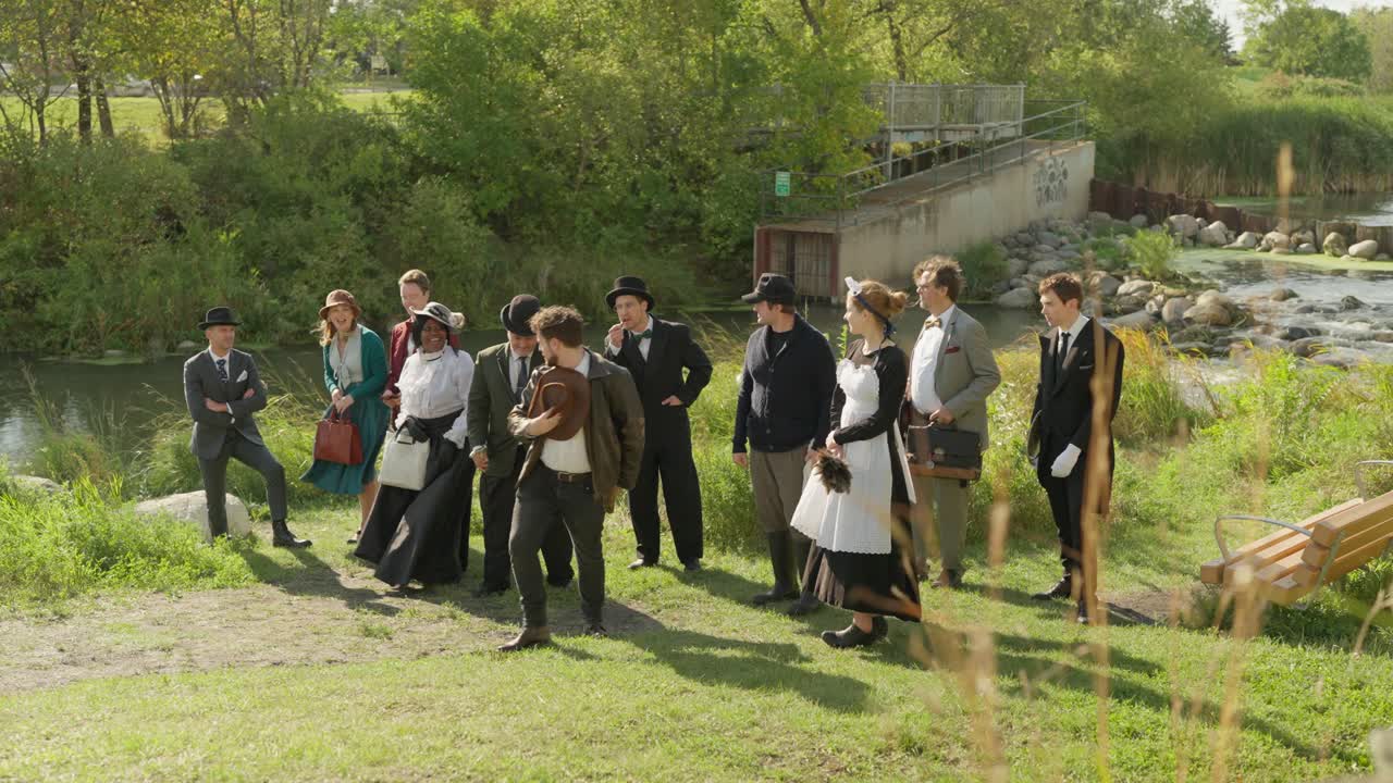 Entire cast of play in vintage attire pose by river dam in sunny park