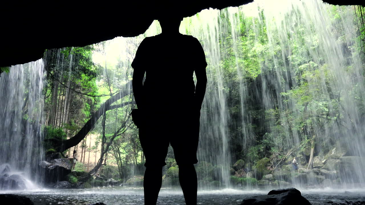 Silhouette of a man standing behind a curtain of water at Nabegataki Falls Kumamoto waterfall in a forest in Japan