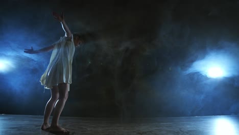 a young female ballerina barefoot jumps on stage and moves in slow motion in a loose white dress