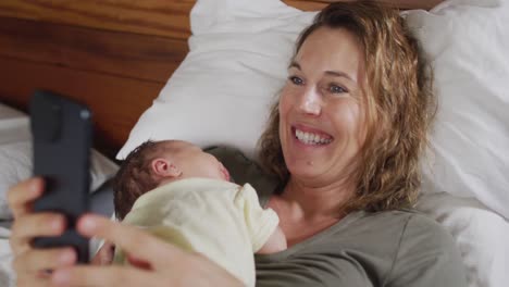 video of happy caucasian mother lying on bed with sleeping newborn baby and using smartphone