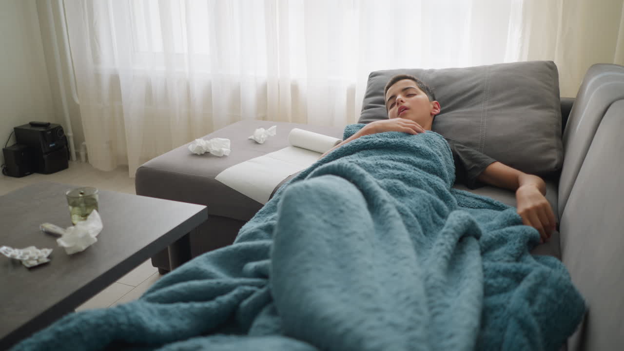 Bambino malato a riposo sul divano, che si copre la bocca mentre tossisce, sistema la coperta per stare più comodo, fazzoletto usato, termometro e tazza di vetro sul tavolo, si riposa mentre non si sente bene