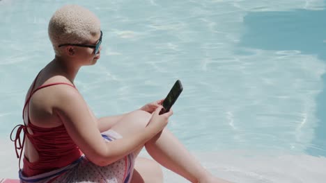 mujer biracial feliz usando un teléfono inteligente y sentada en la piscina en cámara lenta