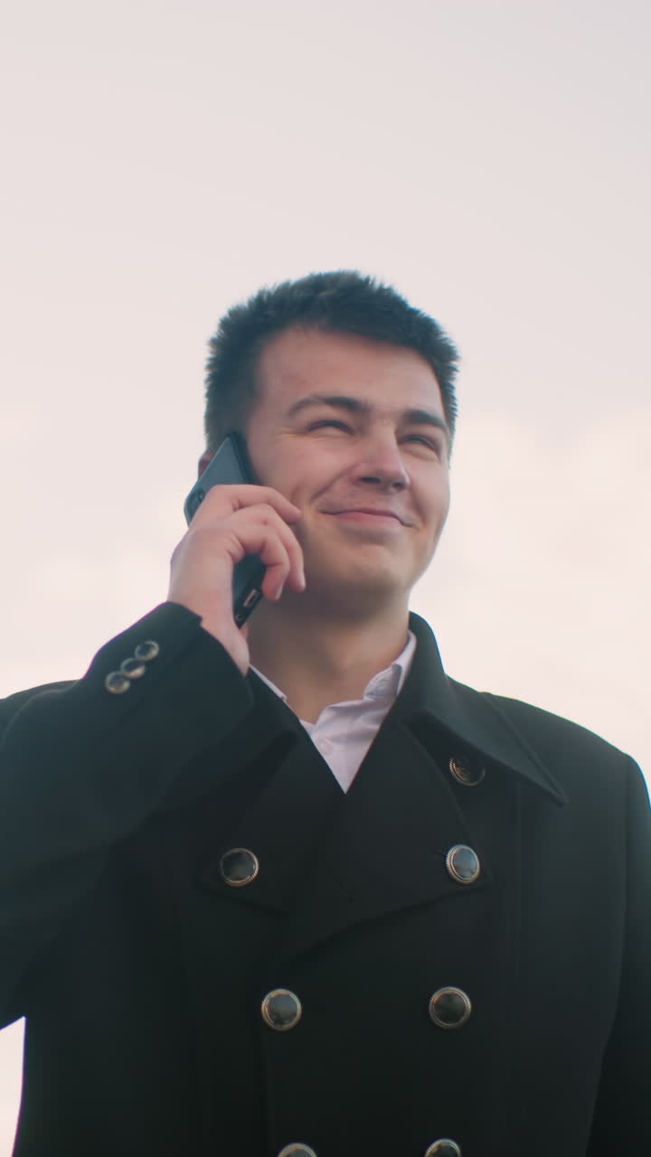 현대적인 유리 건물, 부드러운 빛, 그리고 즐거운 분위기가 있는 활기찬 도시 지역에서 웃고 흥분한 손짓을 하면서 전화로 이야기하는 검은 코트를 입은 남자