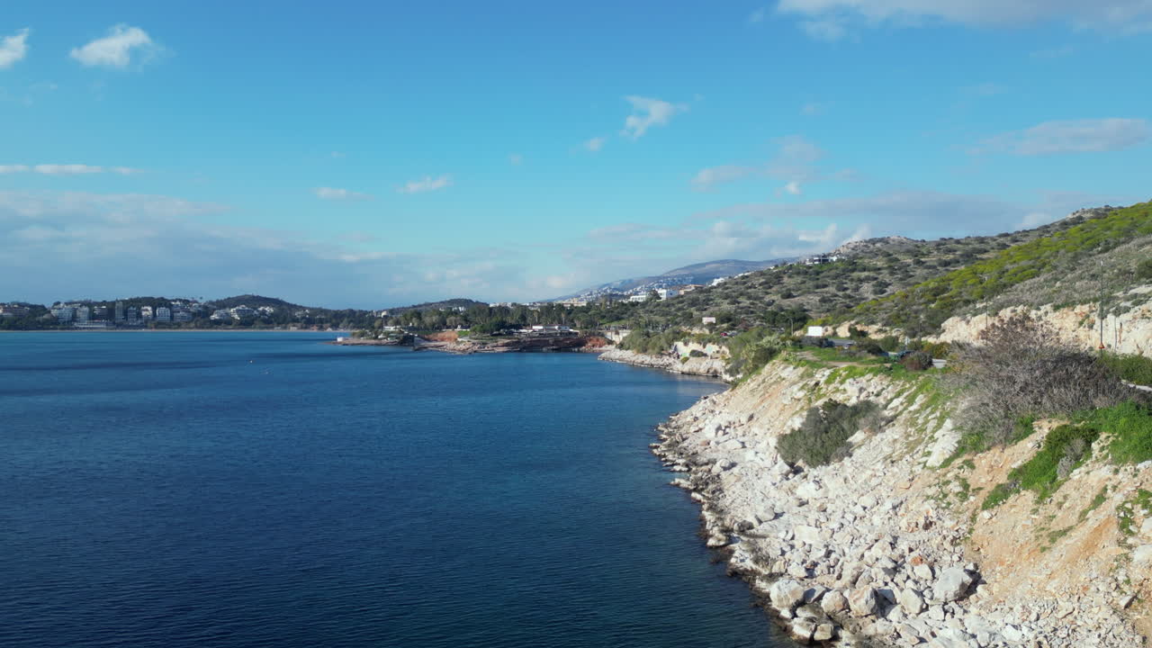 Fly over Athens stunning coastline with the sea on the right and a scenic road on the left. Experience Athens in February, with sunny weather and picturesque clouds dotting the sky.