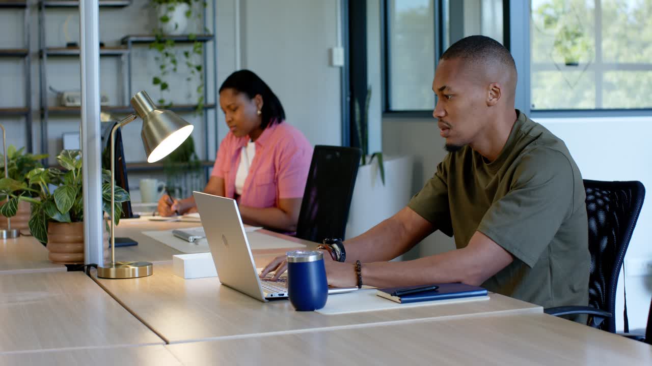Coworkers sitting at table in office, man in green shirt typing on laptop, woman writing notes