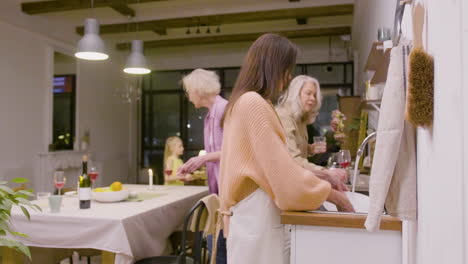 vista lateral de una mujer lavando los platos de la cena familiar en el fregadero de la cocina mientras dos mujeres maduras quitan los platos de la mesa