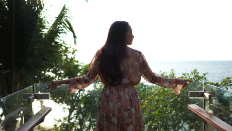 A-young-fit-Asian-woman-in-a-dress,-stands-on-a-glassed-in-balcony-looking-out-at-the-ocean-horizon