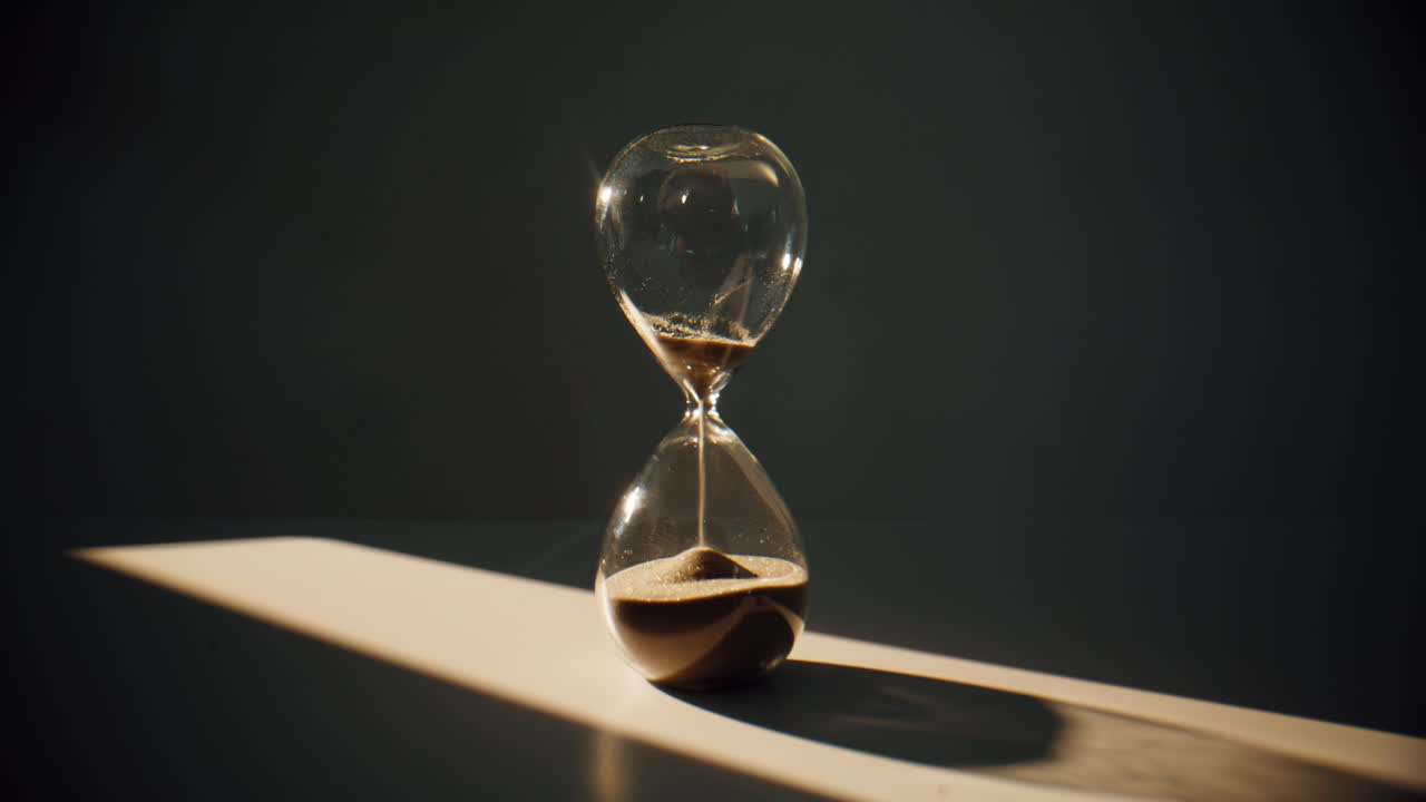Sand flowing inside hourglass standing on surface in evening shadow closeup