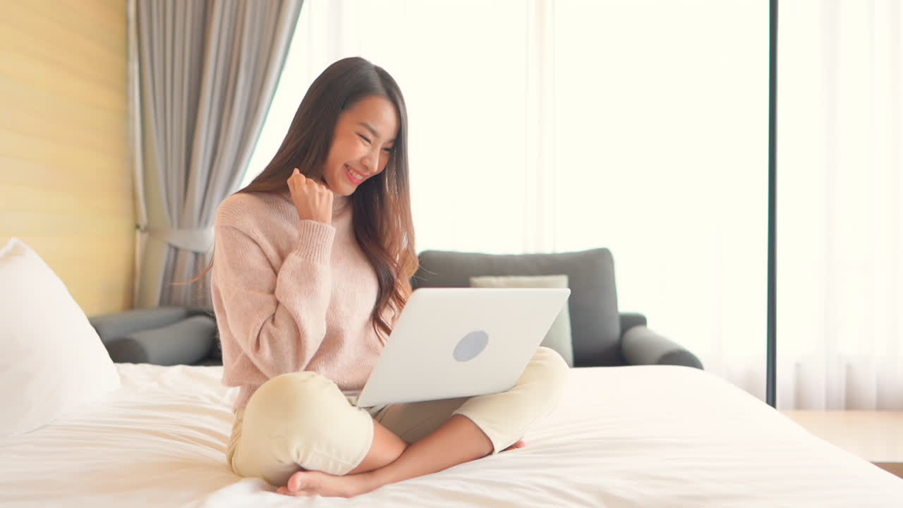 Young Asian woman on hotel bed with laptop press keys, is pleased with results