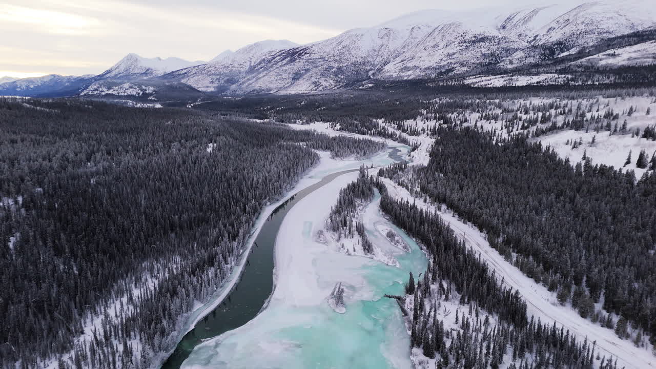 A cinematic aerial pull-out with a tilt-up reveal captures the frozen Takhini River and Kusawa Lake amid fir trees and mountains, epitomizing Yukon’s pristine winter wilderness.