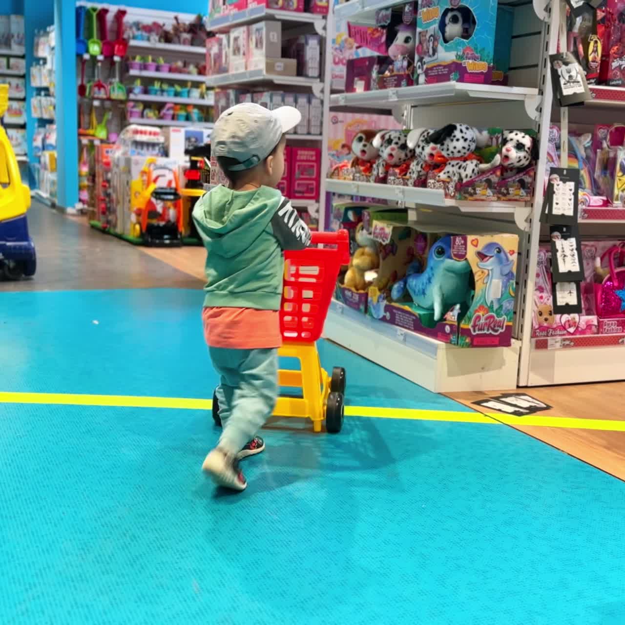 Happy Caucasian baby boy pushes a shopping cart by the mall. Kid choosing a new toy at toy shop