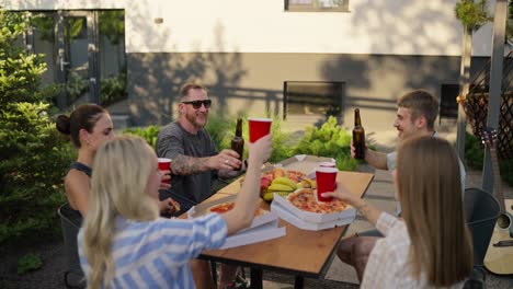 A-happy-company-holds-glasses-and-brown-bottles-in-their-hands-and-clinks-glasses-during-a-shared-lunch-at-a-table-in-the-courtyard-of-a-country-house-on-the-weekend