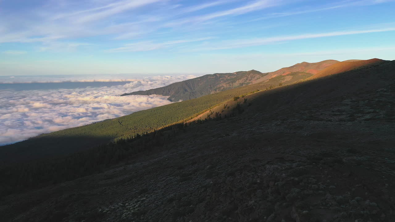 View from the Pico de Teide mountain on a dense cloud inversion below the hills lit by the evening sunlight and the Atlantic ocean visible on the horizon. Cloud inversion below mountains at sunset 4K.
