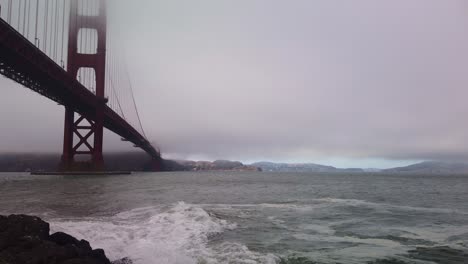 gimbal static shot of rough waves hitting the rocky shoreline underneath the golden gate bridge on a foggy day in san francisco, california