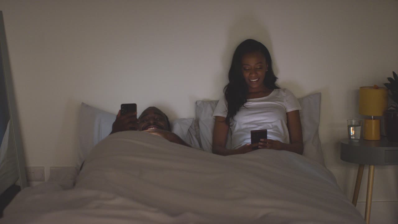 Couple At Home At Night Both Looking At Their Mobile Phones In Bed