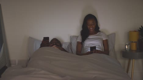 Una-Pareja-En-Casa-Por-La-Noche,-Ambos-Mirando-Sus-Teléfonos-Móviles-En-La-Cama