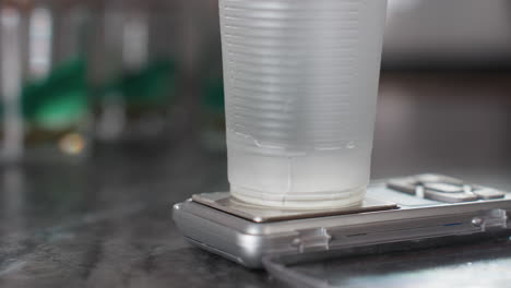 close up disposable cup positioned on kitchen scale as person activates device while pouring liquid substance into cup during ingredient measurement process in modern kitchen with blurred background