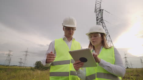 Ein-Elektriker-Und-Eine-Elektrikerin-Auf-Den-Feldern-In-Der-Nähe-Der-Stromübertragungsleitung.-Er-Ist-Elektriker-Und-Leitet-Den-Prozess-Der-Errichtung-Von-Stromleitungen.-Der-Mechaniker-Mit-Helm-Und-Der-Manager-Mit-Tablet