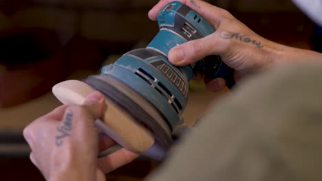 slow motion close-up of person with hand tattoos using electric orbital sanding machine tool to shape a wooden hardwood block spraying dust at home diy garage shed workshop in australia carpentry