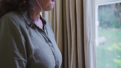 mujer caucásica mayor pensativa y triste sentada en la sala de estar mirando por la ventana