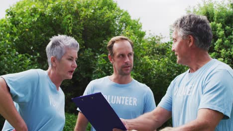 Kaukasisches-älteres-Paar-Mit-Klemmbrett-Und-Mann,-Der-Freiwilligen-T-Shirts-Trägt-Und-Sich-Auf-Dem-Feld-Unterhält