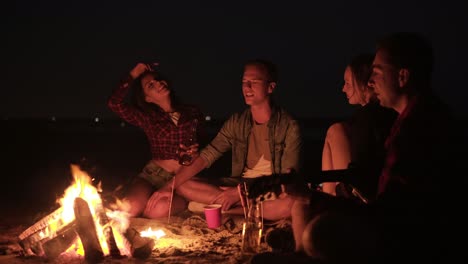 Group-of-friends-hanging-out-together-on-the-beach.-Beautiful-two-couples.-Singing-song-with-guitar,-gesturing.-Friendship-concept.-Bonfire-at-night