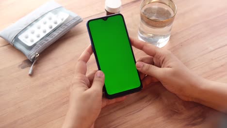 person holding a green screen smartphone on a table with medicine and water