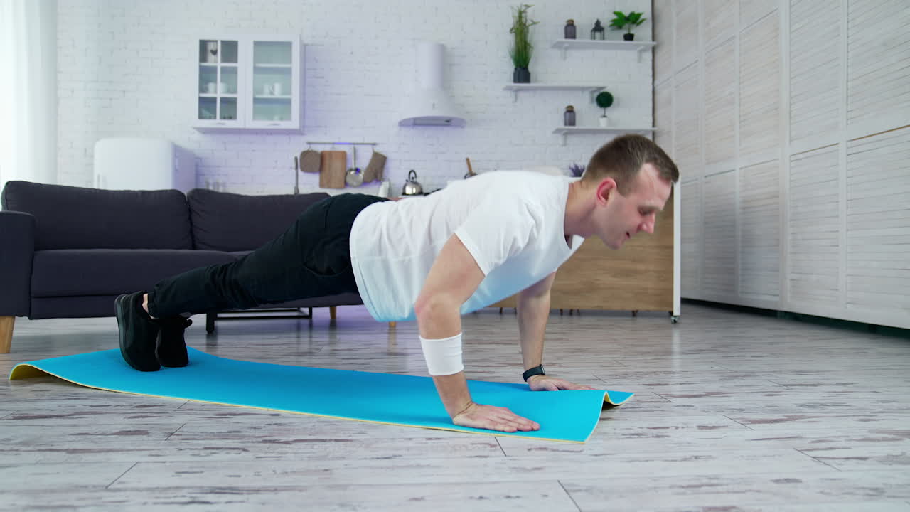 Sportsman training at home. Young man in white t-shirt and black pants exercising on a mat in the room. Side view. Sport during lockdown.