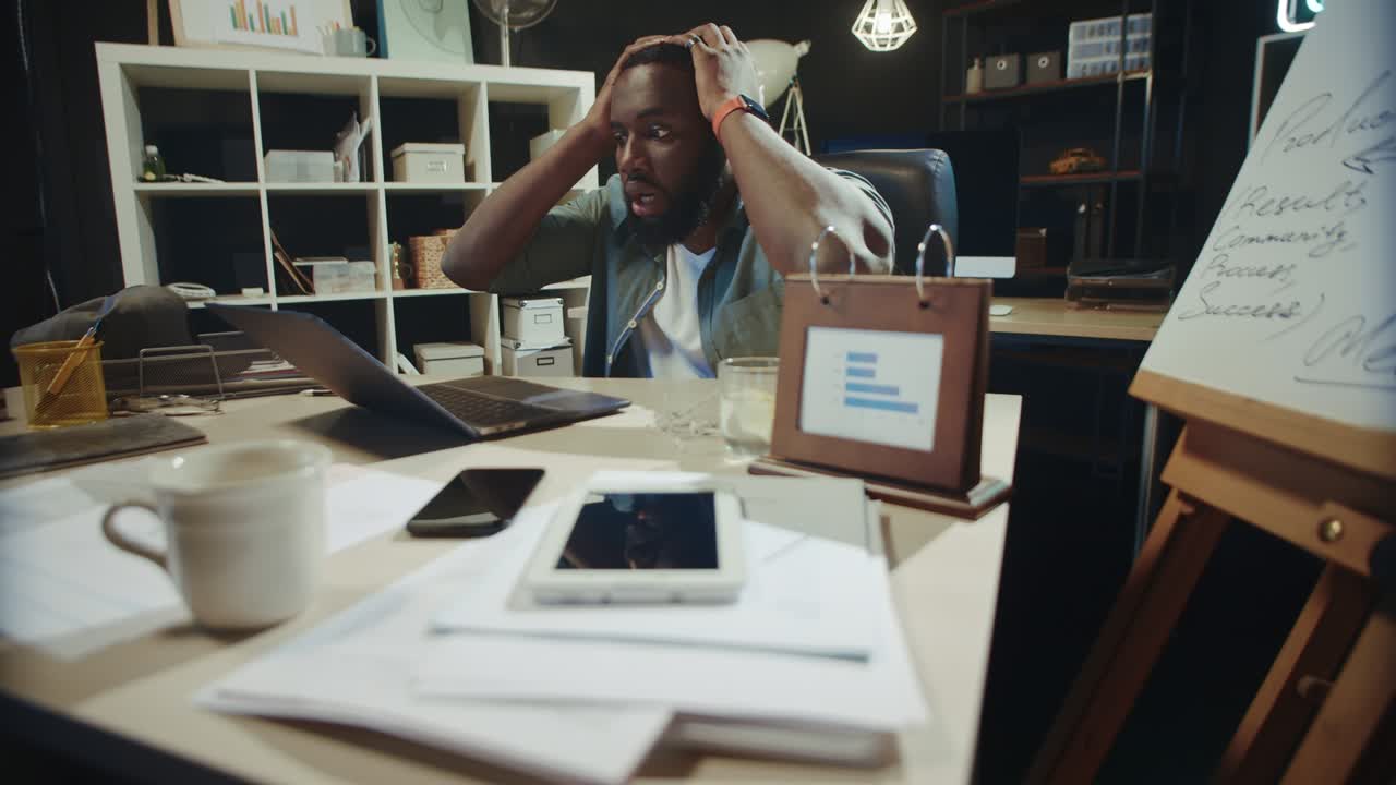 Frustrated african businessman grabbing head in dark coworking.