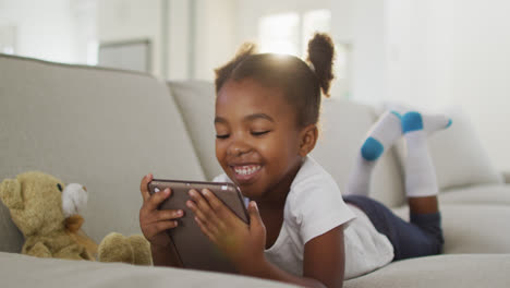 happy african american girl lying on sofa using digital tablet at home