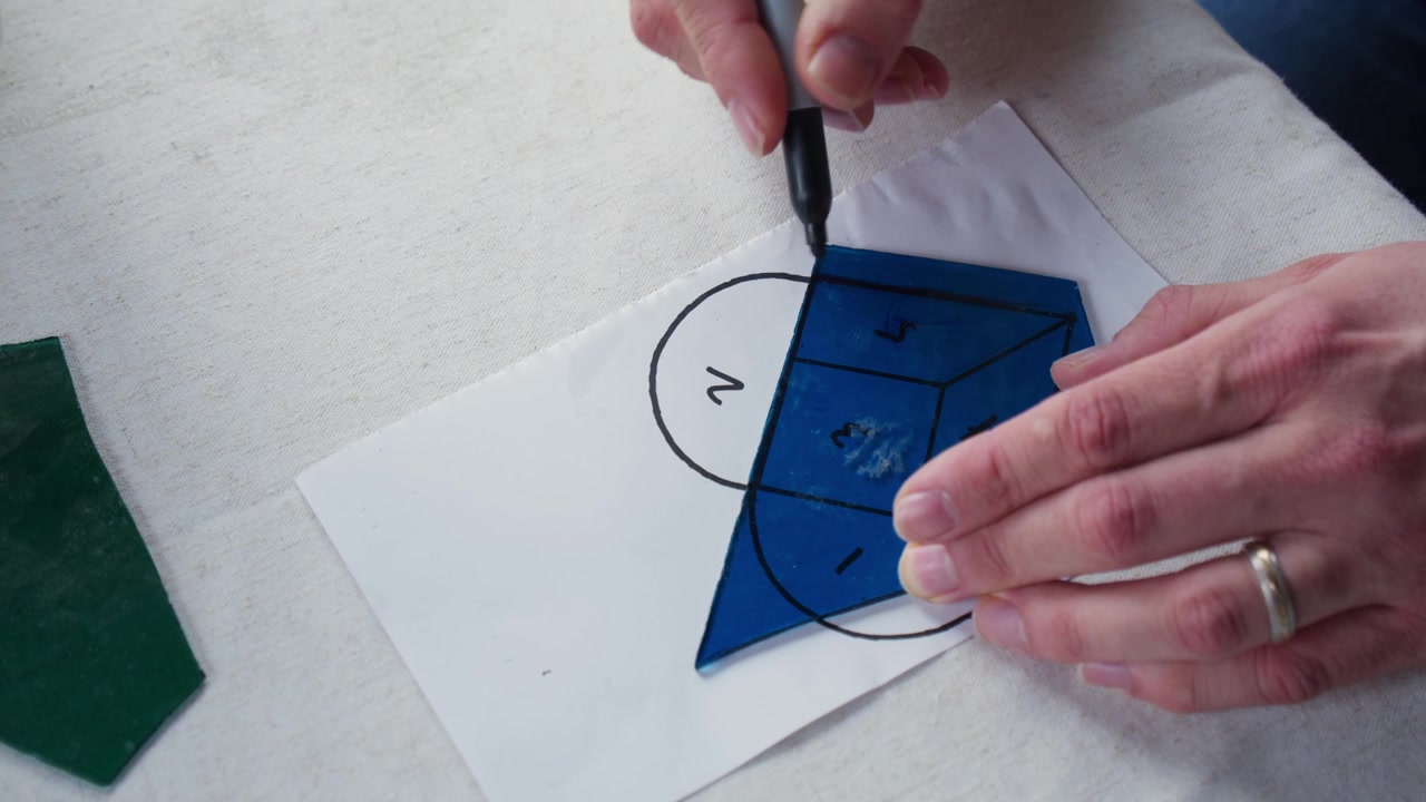 A man wearing a wedding band traces a piece of blue stained glass over one section of a heart template. He is in art class and using a sharpie to outline where he will cut the glass.