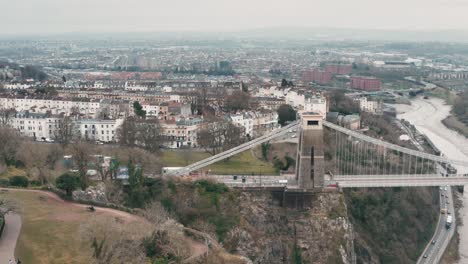 Aerial-shot-of-the-Clifton-suspension-bridge-tracking-from-left-to-right-from-the-observatory-across-the-River-Avon,-Bristol,-during-overcast-cloudy-day