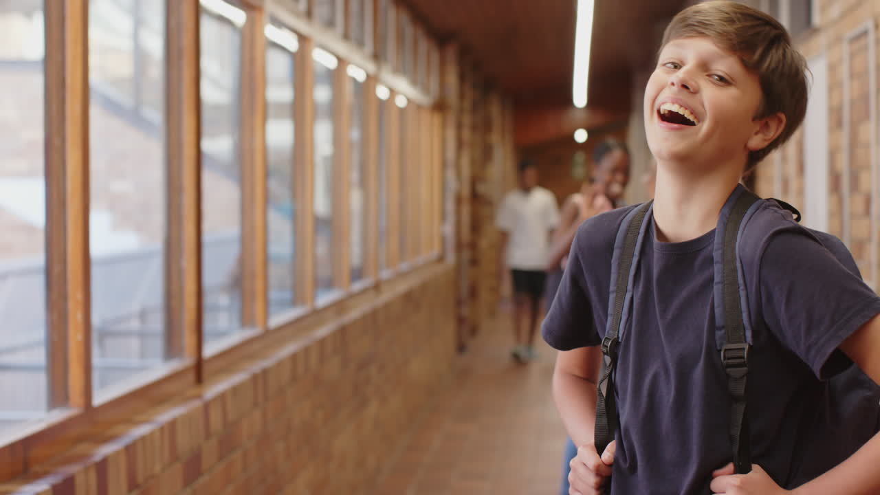 Smiling boy with backpack standing in school hallway with classmates in background