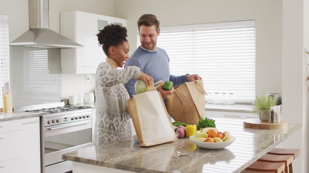video de una feliz pareja diversa desempaquetando comestibles en la cocina