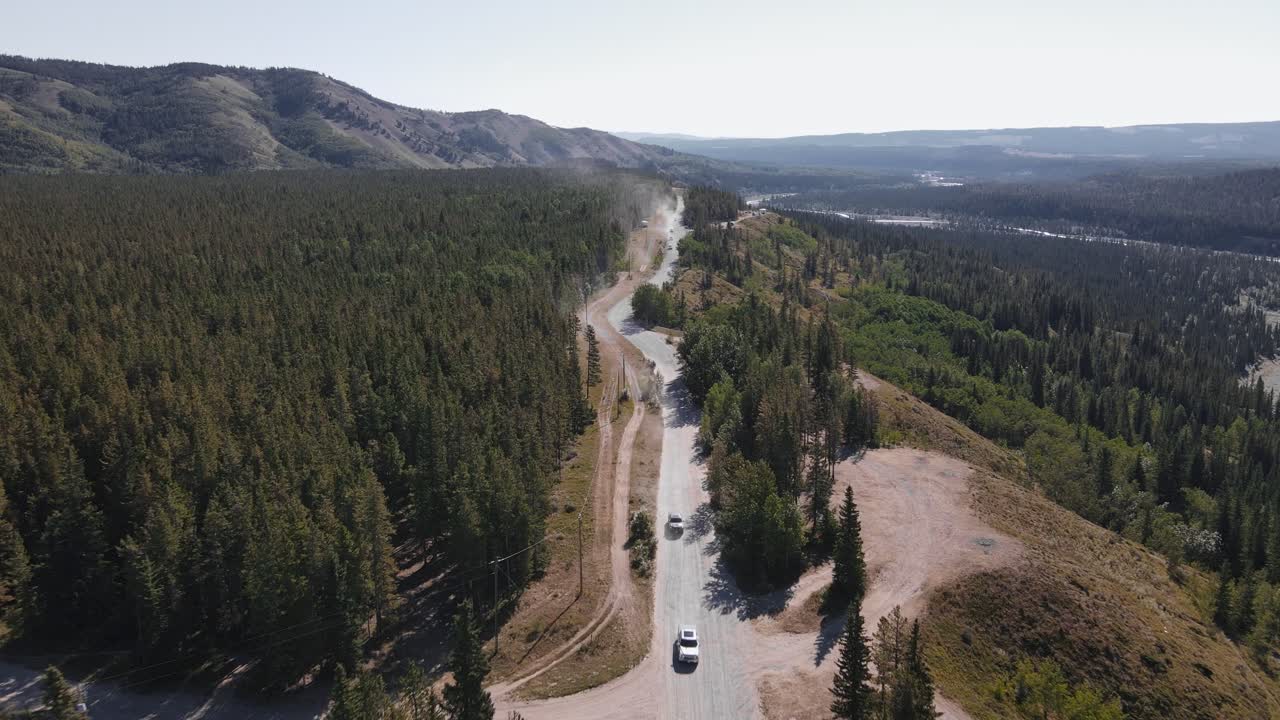 varios coches conduciendo a lo largo de un camino de tala polvoriento que conduce a través de bosques boreales en las afueras de las montañas rocosas